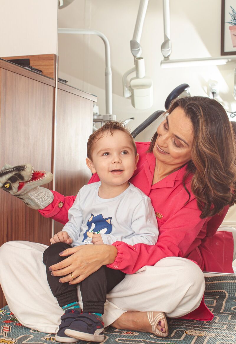 Dra. Viviane sentada no tapete com um bebê no colo, brincando com um fantoche