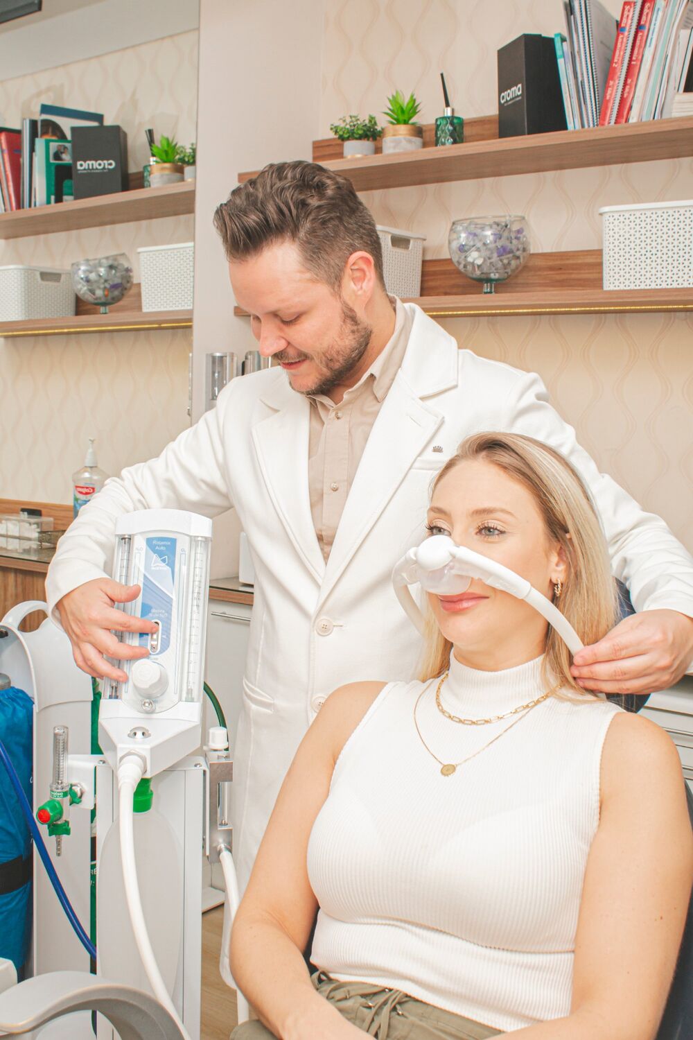 Dr. Marcelo Carvalho ajusta a máscara nasal de óxido nitroso em uma paciente sorrindo, ao lado do equipamento de sedação